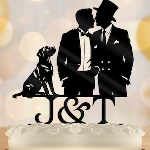 Puede incluir: Adorno para tarta de silueta negra con dos hombres en esmoquin besándose, un perro y las letras "J & T". El adorno se coloca sobre una tarta blanca con un borde de encaje decorativo. El fondo está borroso.