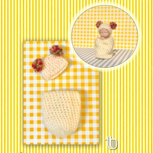 May include: A crocheted baby doll in a cream-colored outfit, including a hat with red and cream pom-poms. The doll is shown in a circular inset and with the hat and a swaddling blanket on a yellow and white checkered surface. The background is yellow and white striped.