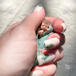 Op de afbeelding: Een hand met witte nagellak met groene bladeren ontwerpen houdt een kleine pop gewikkeld in een groene en witte bloemenstof.