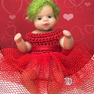 May include: A miniature doll with green hair and a red crocheted dress. The doll has a fair complexion and is posed against a red background with white heart shapes. The doll's dress has a layered skirt made of a red mesh material.