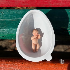 Op de afbeelding: Een klein, wit, eivormig bad van keramiek met een babypop erin. De pop is gemaakt van porselein en heeft een lichtbruine huidskleur. De pop ligt op zijn rug met zijn armen gekruist over zijn borst.