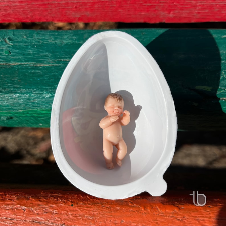 Op de afbeelding: Een klein, wit, eivormig bad van keramiek met een babypop erin. De pop is gemaakt van porselein en heeft een lichtbruine huidskleur. De pop ligt op zijn rug met zijn armen gekruist over zijn borst.