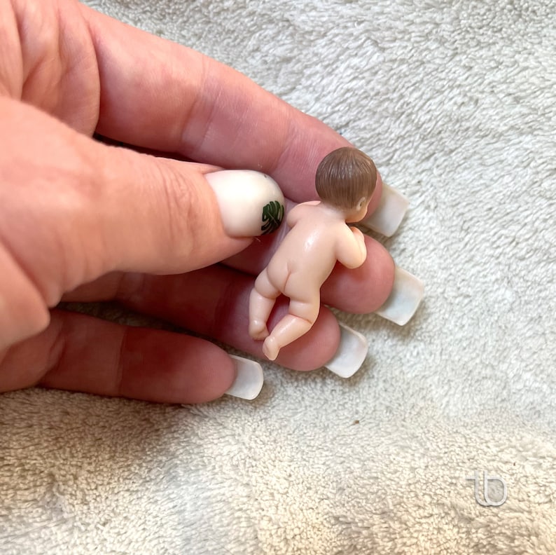 Op de afbeelding: Een kleine, naakte porseleinen pop met bruin haar, liggend op zijn buik met gebogen armen. De pop is ongeveer 5 cm lang.