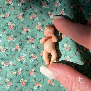 Op de afbeelding: Een kleine, naakte babypop met geschilderd bruin haar en gesloten ogen. De pop ligt op een groene stof met een bloemenpatroon. De pop draagt ​​een witte luier.