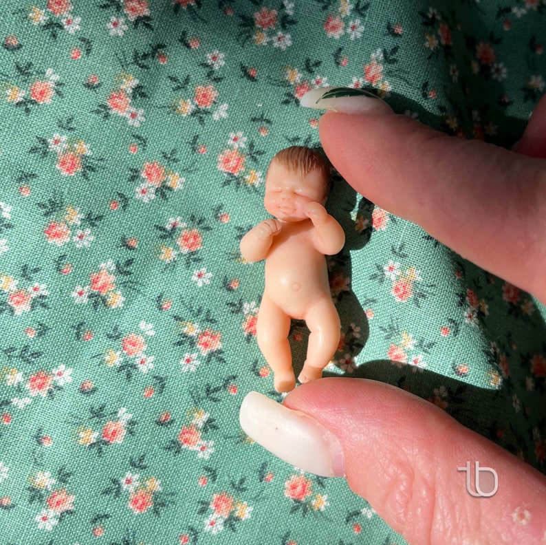 Op de afbeelding: Een kleine, naakte babypop met geschilderd bruin haar en gesloten ogen. De pop ligt op een groene stof met een bloemenpatroon. De pop draagt ​​een witte luier.