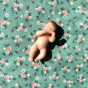Op de afbeelding: Een kleine, naakte babypop met gesloten ogen en een lieve uitdrukking, liggend op een groene stof met een bloemenpatroon.