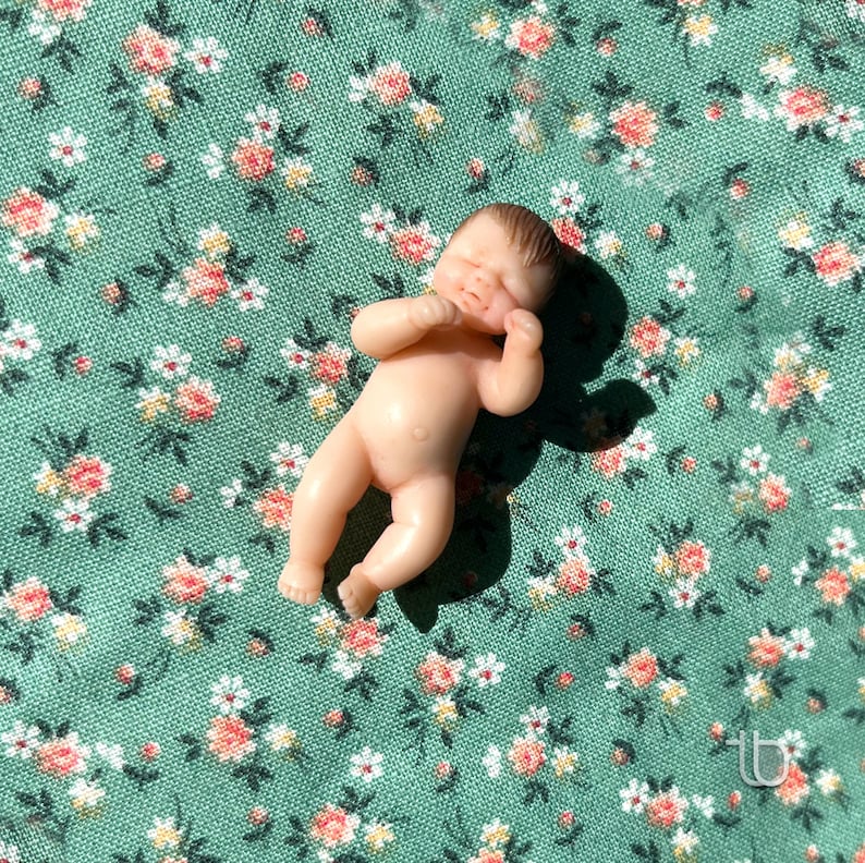 Op de afbeelding: Een kleine, naakte babypop met gesloten ogen en een lieve uitdrukking, liggend op een groene stof met een bloemenpatroon.