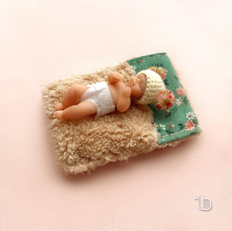 Op de afbeelding: Een kleine babypop met een witte gehaakte muts en een luier, liggend op een bruin, pluche deken met een groene, bloemige stoffen rand.