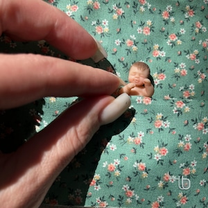 Op de afbeelding: Een kleine, slapende babypop van porselein ligt op een groene, bloemige stof. De pop heeft een lichtroze teint en draagt een witte luier.