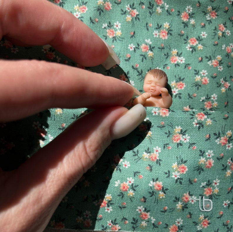 Op de afbeelding: Een kleine, slapende babypop van porselein ligt op een groene, bloemige stof. De pop heeft een lichtroze teint en draagt een witte luier.