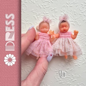 May include: Two miniature baby dolls wearing pink dresses and headbands with bows. The dolls are held by a hand with a light purple nail. The background is a white embossed floral pattern. The word "DRESS" is printed vertically on the left side of the image.