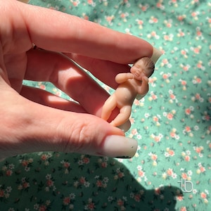 Op de afbeelding: Een kleine, realistische babypop van siliconen, met beschilderde details, wordt in een hand gehouden. De pop ligt in slaaphouding met licht gebogen armen en gesloten ogen.