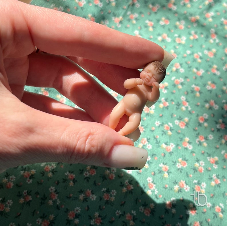 Op de afbeelding: Een kleine, realistische babypop van siliconen, met beschilderde details, wordt in een hand gehouden. De pop ligt in slaaphouding met licht gebogen armen en gesloten ogen.