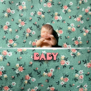 Op de afbeelding: Een kleine, realistische babypop die op een groene stof met bloemenpatroon ligt. De pop heeft een lieve uitdrukking en draagt een witte luier. Het woord "BABY" staat in roze letters met zwarte contouren op de stof.