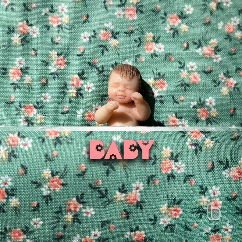Op de afbeelding: Een kleine, realistische babypop die op een groene stof met bloemenpatroon ligt. De pop heeft een lieve uitdrukking en draagt een witte luier. Het woord "BABY" staat in roze letters met zwarte contouren op de stof.