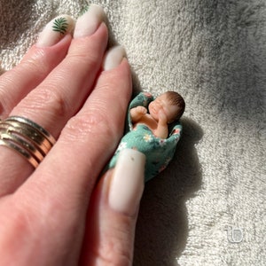 Op de afbeelding: Een kleine, realistische babypop, gewikkeld in een groene en witte bloemenstof, wordt in de palm van een hand gehouden. De pop heeft een lieve, slapende uitdrukking.