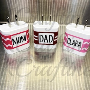 May include: Three white phone chargers with silver prongs. Each charger has a rectangular label with a name: "MOM" in a red and white chevron pattern, "DAD" in a maroon and white striped pattern, and "CLARA" in a pink and white chevron pattern.