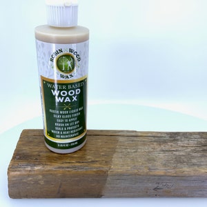 Puede incluir: Una botella de cera para madera a base de agua Robin Wood se encuentra junto a una tabla de madera envejecida. La botella de cera es blanca y verde, con el texto "Rustic Wood Liquid Wax" y "Silky Gloss Finish". La tabla de madera est&aacute; parcialmente tratada.