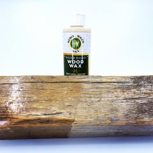 Puede incluir: Una botella de cera para madera a base de agua Robin Wood, con tapa blanca y etiqueta con dise&ntilde;o verde y dorado. La botella se encuentra sobre una tabla de madera envejecida, con una secci&oacute;n tratada que muestra el efecto de la cera. El producto est&aacute; etiquetado como cera l&iacute;quida para madera r&uacute;stica.