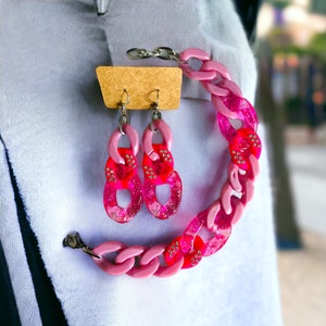 May include: A pink and red chunky chain necklace and matching earrings. The necklace and earrings are made of plastic and have a sparkly finish.
