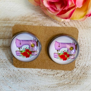 Könnte beinhalten: Ein Paar silberfarbene Ohrringe mit einem Design aus einer rosa Nähmaschine und roten Rosen auf einer braunen Karte.