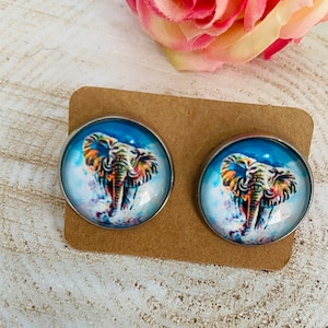 Könnte beinhalten: Ein Paar silberne Ohrstecker mit bunten Elefantenmotiven. Die Elefanten sind von einem blauen und weißen Aquarellhintergrund umgeben.