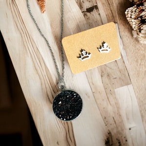 Puede incluir: Un collar de cadena de plata con un colgante redondo negro con una superficie texturizada brillante. El collar se combina con un juego de pendientes de plata con forma de corona con una sola piedra transparente en el centro.