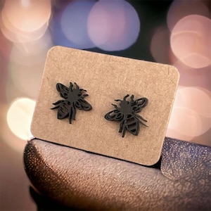 Könnte beinhalten: Ein Paar schwarze, bienenförmige Ohrringe, präsentiert auf einer kleinen, rechteckigen, braunen Karte. Die Ohrringe sind ausgeschnittene Designs, die die Details der Flügel und des Körpers der Biene zeigen. Der Hintergrund ist verschwommen mit weichen, farbenfrohen Bokeh-Lichtern.