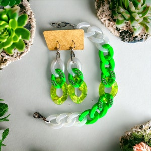 May include: A green and white chain necklace and matching earrings. The necklace and earrings are made of plastic and have a chunky chain design. The earrings are dangle earrings.