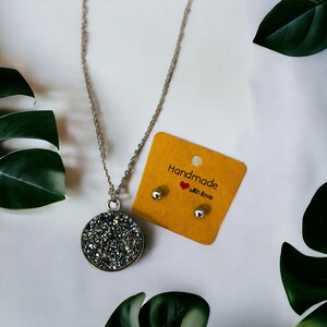 Puede incluir: Un collar de plata con un colgante redondo cubierto de purpurina plateada brillante. El colgante está unido a una delicada cadena de plata. El collar viene con un par de pendientes de botón de plata a juego. La tarjeta dice "Handmade with love".