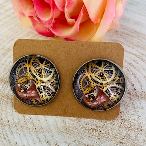 Könnte beinhalten: Ein Paar runde, silberfarbene Ohrringe mit Steampunk-Design. Die Ohrringe zeigen ein kreisförmiges Bild von goldenen Zahnrädern und einem braunen, stilisierten Horn.