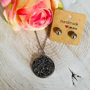 Puede incluir: Un collar de plata con un colgante redondo cubierto de purpurina negra brillante. El colgante cuelga de una cadena de plata. El collar se muestra sobre una superficie de madera con un par de pendientes de botón de plata y una tarjeta que dice "Hecho a mano con amor".