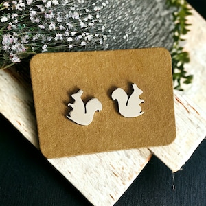 Puede incluir: Un par de pendientes de botón de color beige con forma de ardilla sobre una tarjeta marrón.