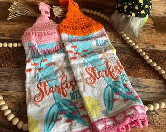 May include: Two crocheted dish towels with a mermaid and starfish design. The towels are pink and orange with white pom-poms. The towels have the text "Mermaid Kisses & Starfish Wishes".