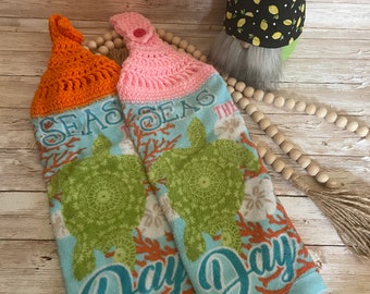 May include: Two crocheted dish towels with a sea turtle and coral design. The towels have the text "Seas the Day" on them. The towels are hanging on a wooden surface.