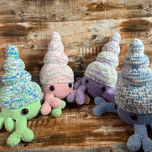 May include: Four crocheted hermit crab plushies in different colors. The crabs are wearing crocheted shells in shades of pink, purple, green, and blue.