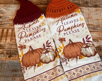 May include: Two white kitchen towels with a fall theme. The towels have a crocheted loop at the top. One towel has a red crocheted loop and says "Autumn Leaves & Pumpkins Please". The other towel has an orange crocheted loop and says "Autumn Leaves & Pumpkins Please".