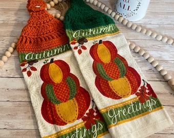 May include: Two kitchen towels with a fall theme. The towels are white with a green and orange crocheted loop at the top. The towels feature a stack of pumpkins with the words "Autumn Greetings" printed on them.