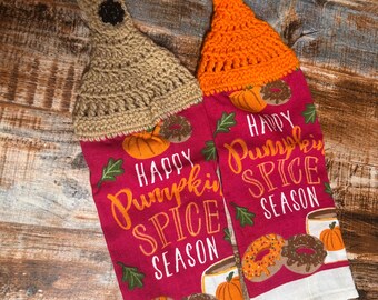 May include: Two kitchen towels with crocheted tops. The towels are red with the words "Happy Pumpkin Spice Season" and feature images of pumpkins, donuts, and coffee cups. One towel has a tan crocheted top, and the other has an orange crocheted top.