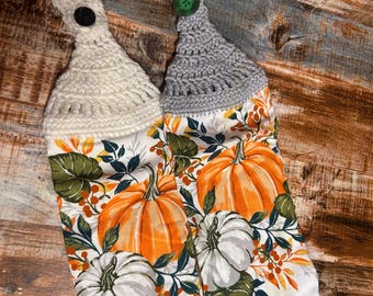 May include: Two hanging kitchen towels with a fall theme. The towels feature a pumpkin print in orange and white, with green leaves and berries. The tops are crocheted, one in cream and the other in gray, each with a button.