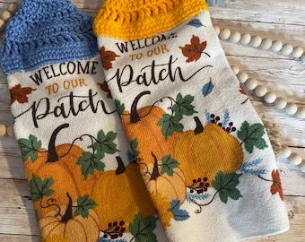 May include: Two white kitchen towels with crocheted tops. The towels feature a fall theme with pumpkins, leaves, and the text "Welcome to our Patch". One towel has a yellow crocheted top and the other has a blue crocheted top.