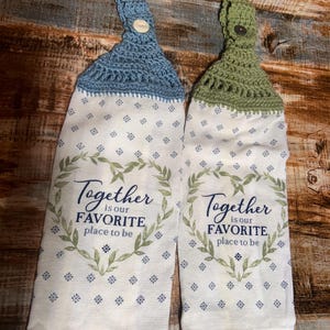 May include: Two hanging kitchen towels with a floral pattern and the text "Together is our favorite place to be." One towel has a blue crocheted top and the other has a green crocheted top. Each towel has a button closure.