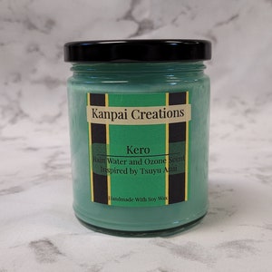 Puede incluir: Un tarro de vela de vidrio verde con tapa negra. La etiqueta dice "Kanpai Creations Kero Rain Water and Ozone Scent Inspired by Tsuyu Asui Handmade With Soy Wax".