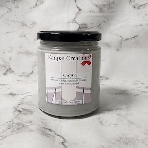 Puede incluir: Un tarro de vela de vidrio gris con tapa negra. La etiqueta dice "Kanpai Creations" y "Vaggie" con la descripción del aroma "Incienso, cedro, pachulí, naranja y clavo". La etiqueta también dice "Vela de soja hecha a mano".