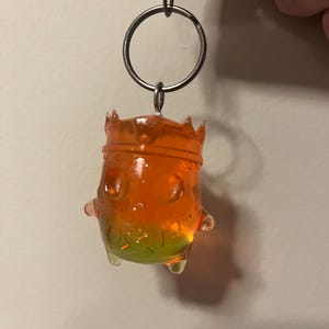 May include: A translucent orange and green keychain featuring a small, rounded character wearing a crown. The figure has a smooth, glossy finish with subtle internal glitter particles, suspended from a silver-toned metal split ring and lobster clasp.