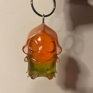 May include: A translucent keychain featuring a small, hooded figure with a smooth, rounded head. The upper portion of the figure is a vibrant orange, transitioning into a bright lime green at the base. It is attached to a circular metal ring.