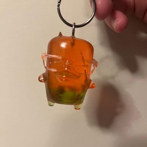 May include: A translucent orange keychain featuring a small, rounded character with two tiny horns and clear plastic glasses. The figure has a gradient effect, transitioning from a vibrant orange at the top to a lime green hue at the base.