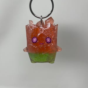 May include: A keychain featuring a small, translucent character figure with a textured, glittery orange top and a solid green base. The figure has two purple circular eyes and a small purple mouth, with jagged, crystal-like protrusions on its head.