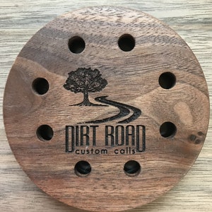 May include: A round wooden call holder with a tree and winding road design. The holder has eight holes for storing calls. The text "Dirt Road Custom Calls" is engraved on the holder.
