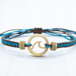 Könnte beinhalten: Ein Makramee-Armband mit einem Wellenmuster. Das Armband hat ein gewebtes Band mit abwechselnd blauen und braunen Fäden. Ein Holzanhänger in Wellenform ist mittig angeordnet, und an jedem Ende befinden sich Holzperlen.